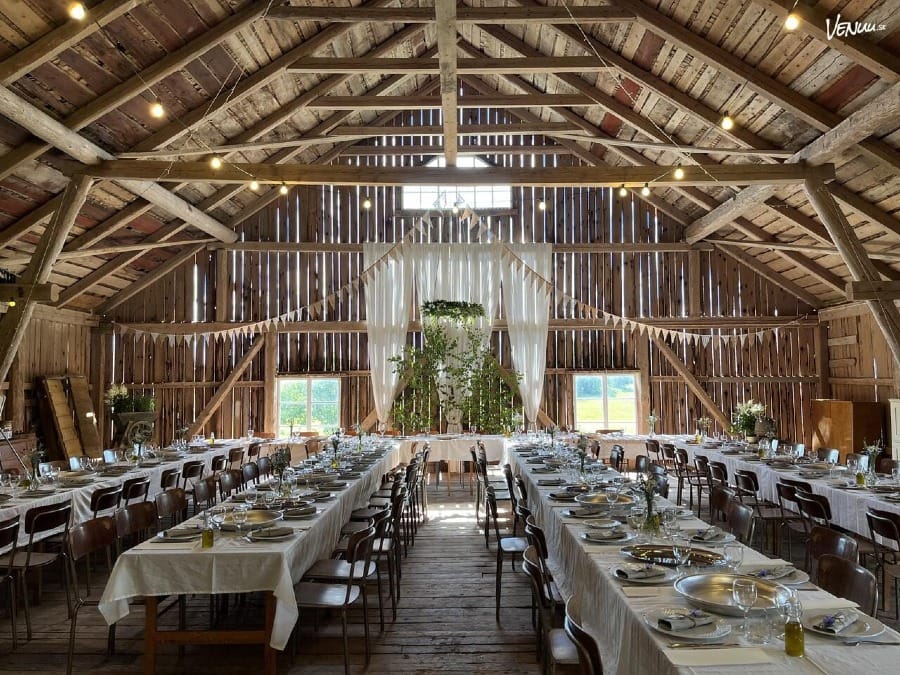 Interiören av en vackert dekorerad lada i Lohärad med långa bord uppställda för en fest eller bröllop. Taket är högt och byggt av träbjälkar, vilket ger en rustik och charmig atmosfär. Borden är dukade med vita dukar, porslin och glas, redo för gästerna.