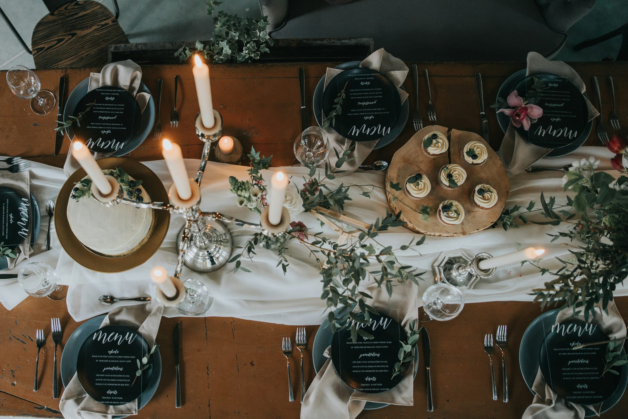 En elegant bröllopsdukning med en rustik touch. Ett långt träbord är dekorerat med ljusa dukar, levande ljus i en silverkandelaber, gröna blad och en vackert dekorerad tårta. Svarta menyer med vit text ligger på tallrikarna, vilket skapar en stilfull kontrast.