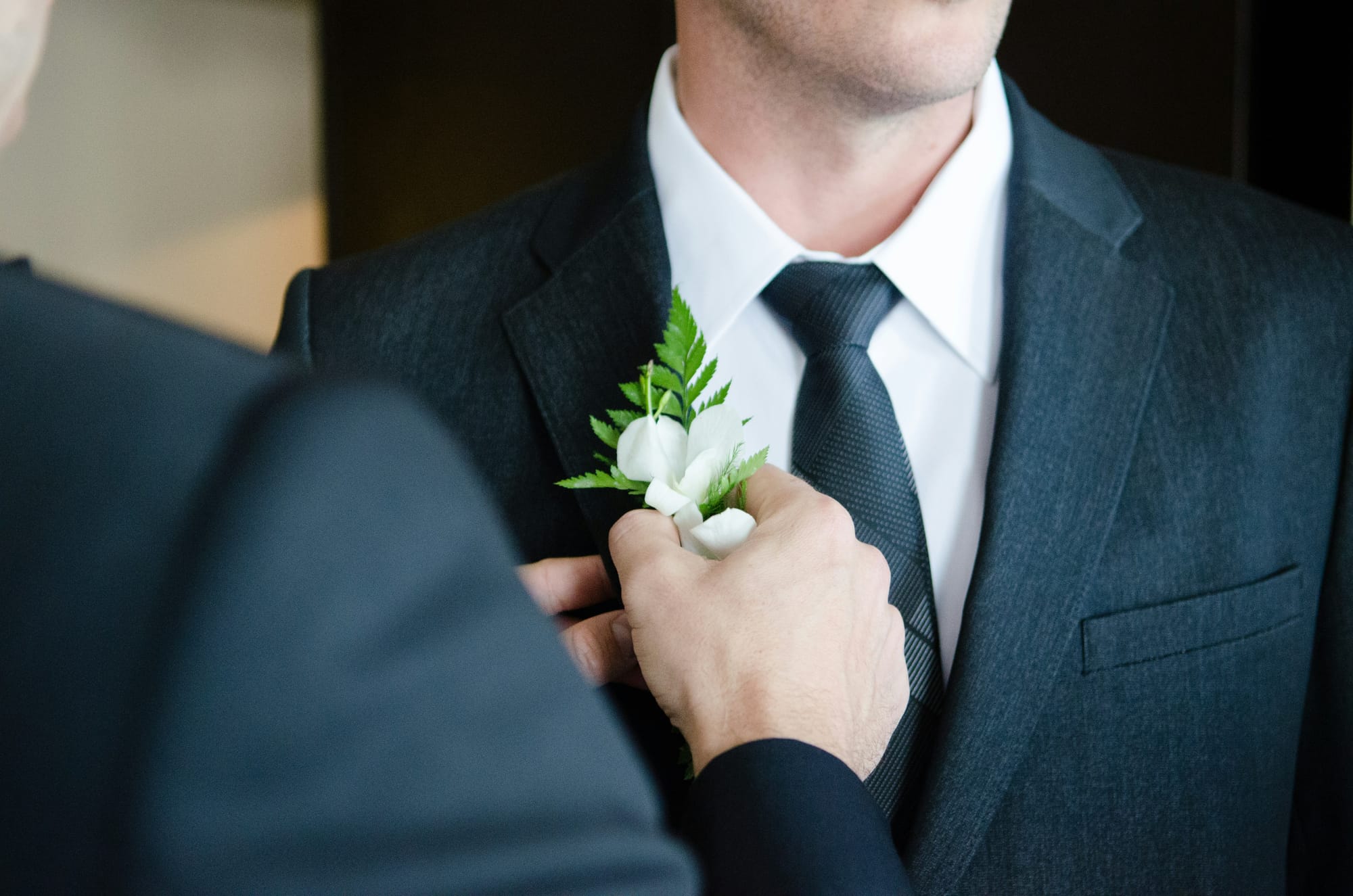 Närbild på en man i en mörk kostym medan någon fäster en boutonnière med vita blommor och gröna blad på hans kavajslag. En stilfull och klassisk detalj inför bröllopsceremonin.