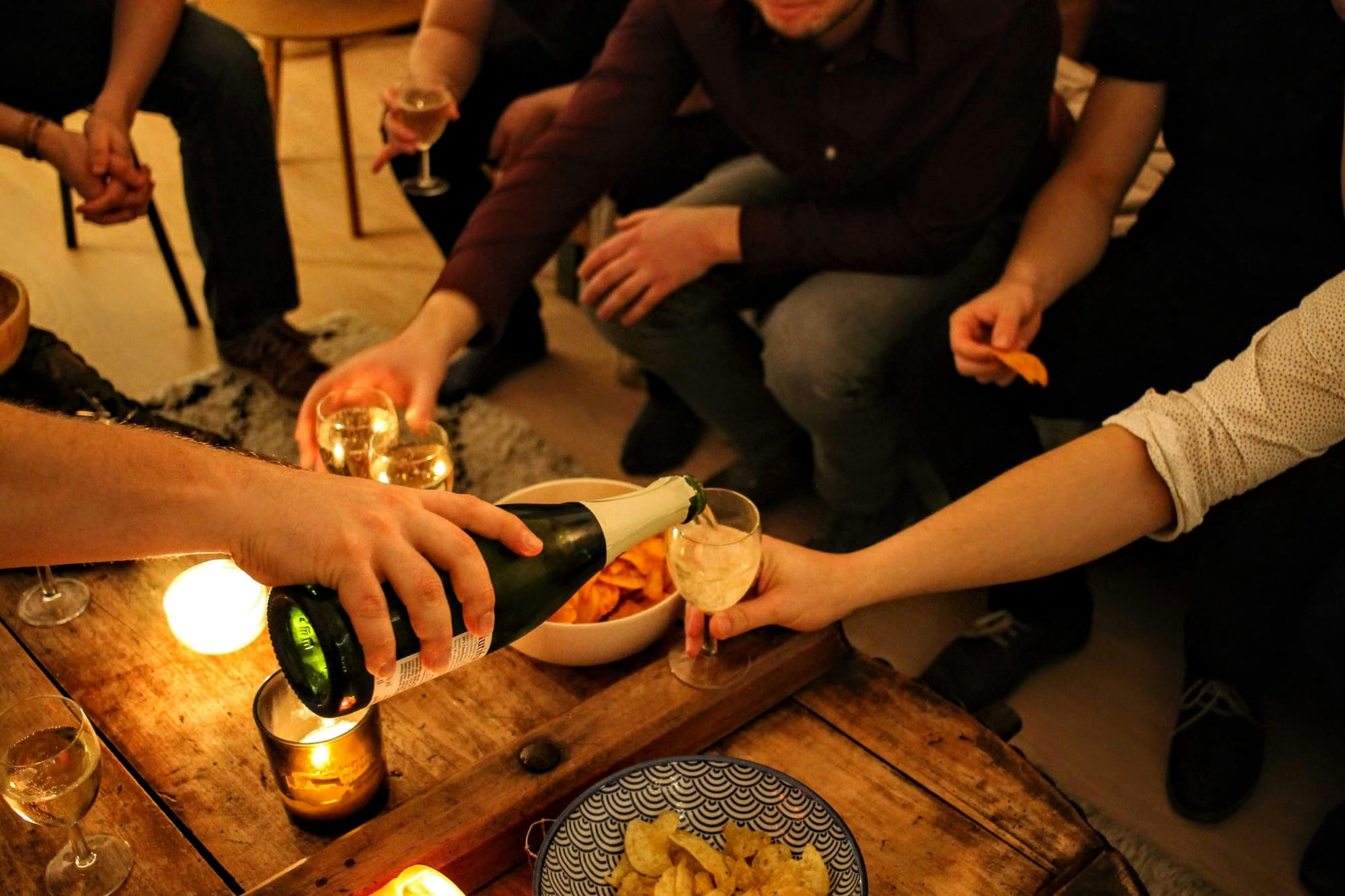 En närbild på händer som spelar kort under en omgång stillasittande vuxenlekar, med flaskor och glas fyllda med dryck på ett träbord.